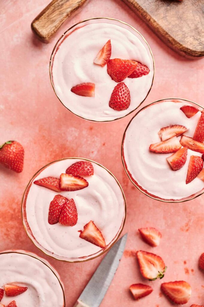 Four glass bowls of Strawberry Mousse are topped with fresh strawberry slices, set on a pink surface alongside a knife, cutting board, and scattered strawberry pieces.