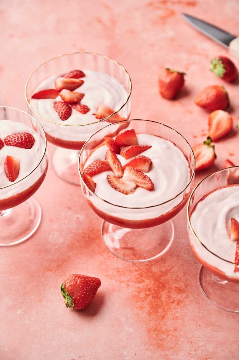 Four glass cups of strawberry mousse, each topped with sliced strawberries, are arranged on a pink surface alongside whole and cut strawberries.