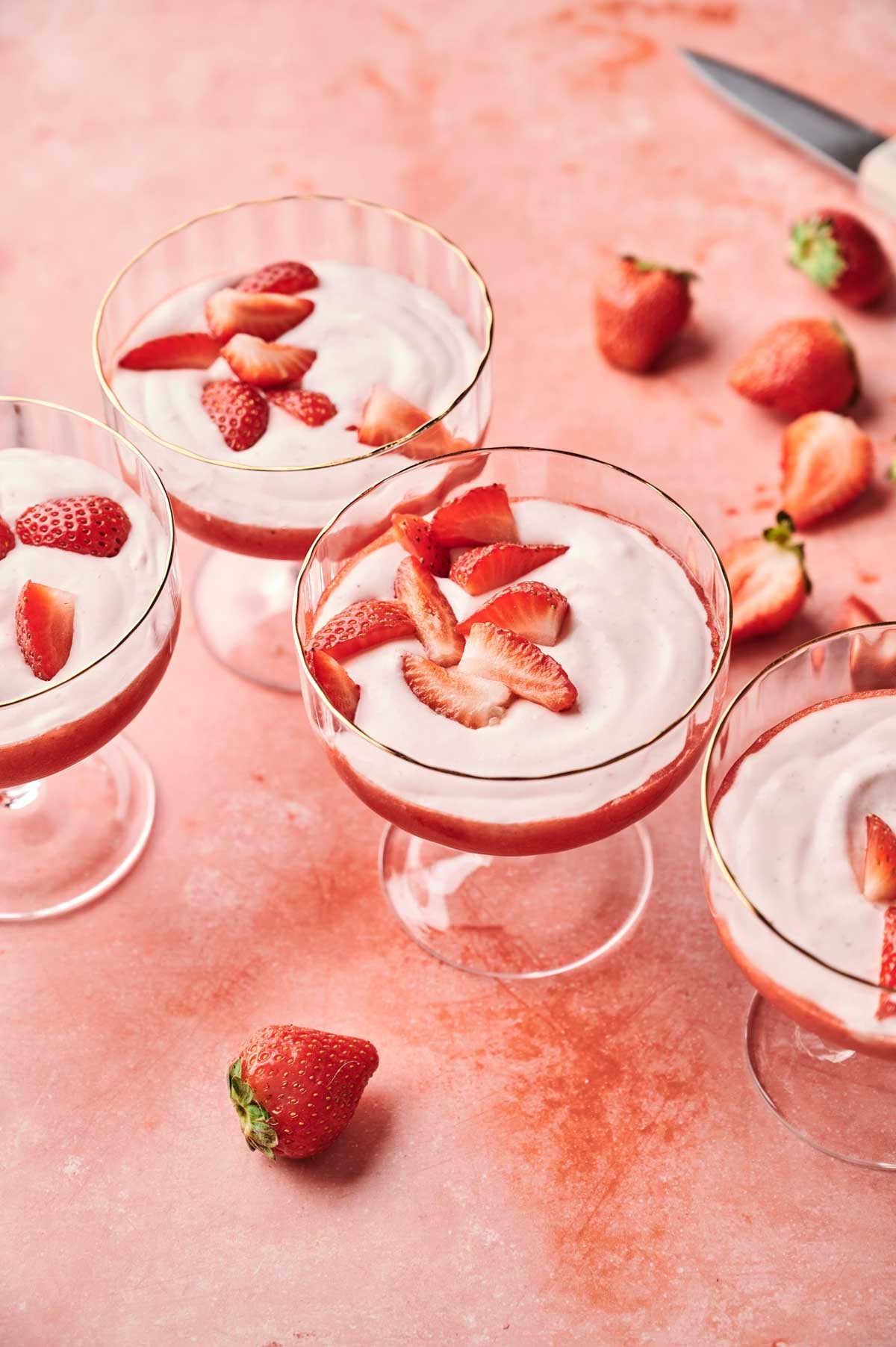 Four glass cups of strawberry mousse, each topped with sliced strawberries, are arranged on a pink surface alongside whole and cut strawberries.
