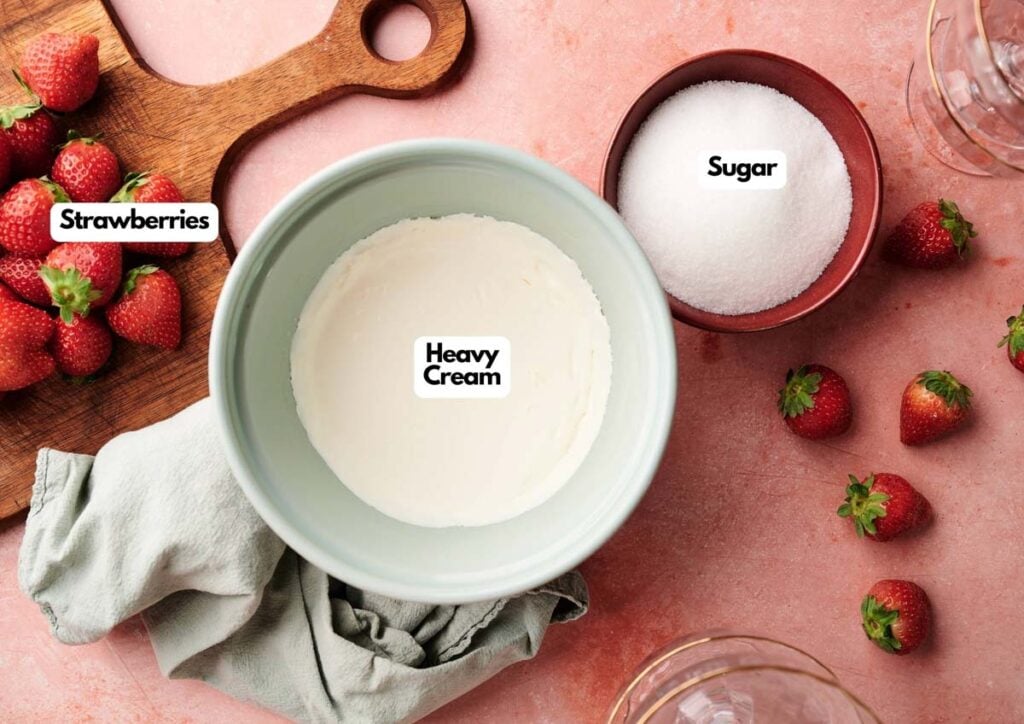 A bowl of heavy cream, a bowl of sugar, and a cutting board with fresh strawberries on a pink surface&mdash;perfect ingredients for making delightful Strawberry Mousse.