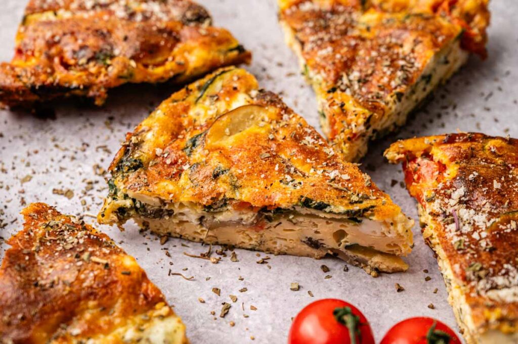 Slices of vegetable frittata with a golden-brown top are arranged on parchment paper, highlighting the colorful layers. Two cherry tomatoes add a vibrant touch beside the delicious Vegetable Frittata.
