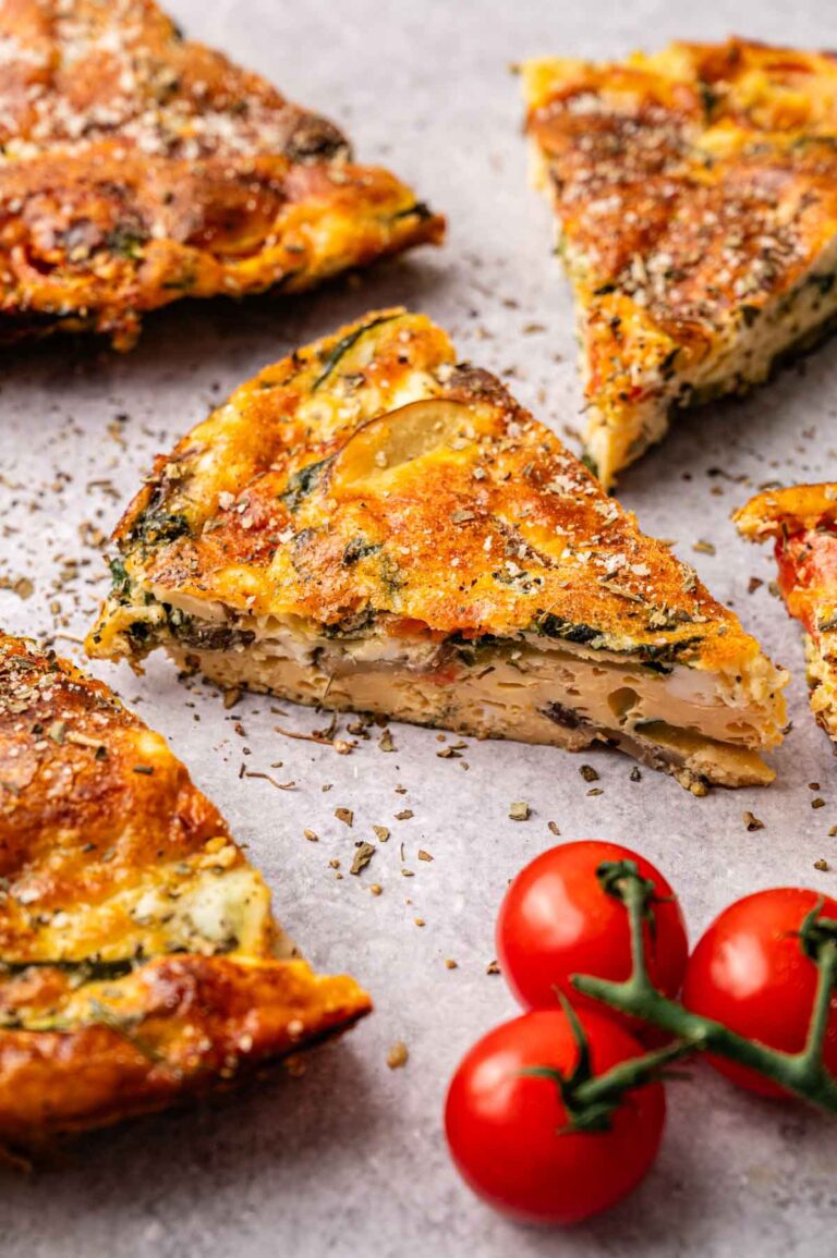 Slices of vegetable frittata are arranged on a light surface with a small vine of cherry tomatoes nearby, highlighting the fresh ingredients in this classic vegetable frittata.