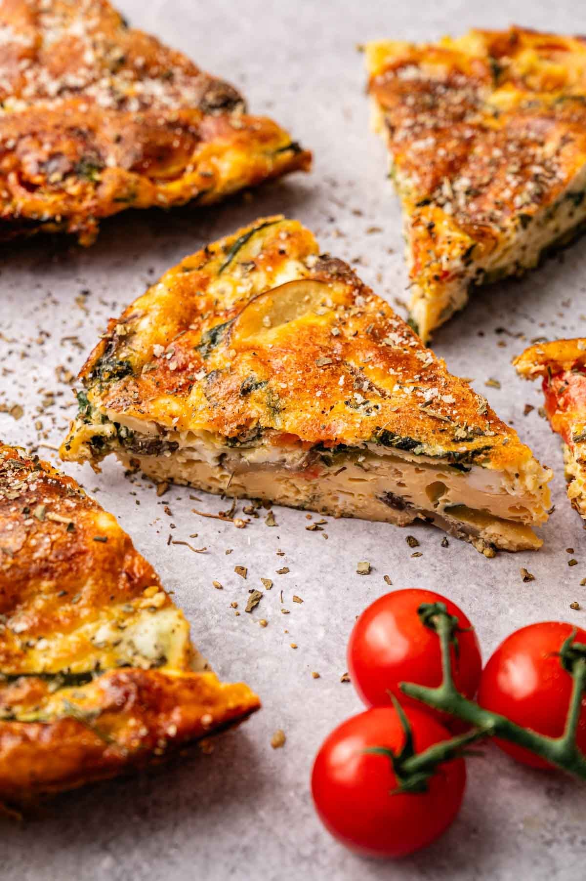 Slices of vegetable frittata are arranged on a light surface with a small vine of cherry tomatoes nearby, highlighting the fresh ingredients in this classic vegetable frittata.