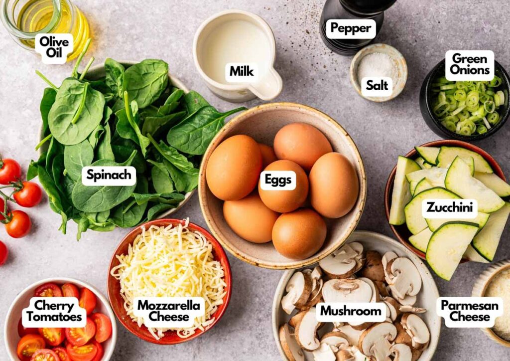 A variety of Vegetable Frittata ingredients are arranged on a table, including eggs, spinach, zucchini, mushrooms, cheese, cherry tomatoes, green onions, olive oil, milk, salt, and pepper.