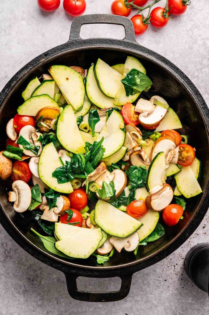 A cast iron pan filled with sliced zucchini, cherry tomatoes, mushrooms, and spinach sits on a light surface, ready to be transformed into a flavorful Vegetable Frittata.