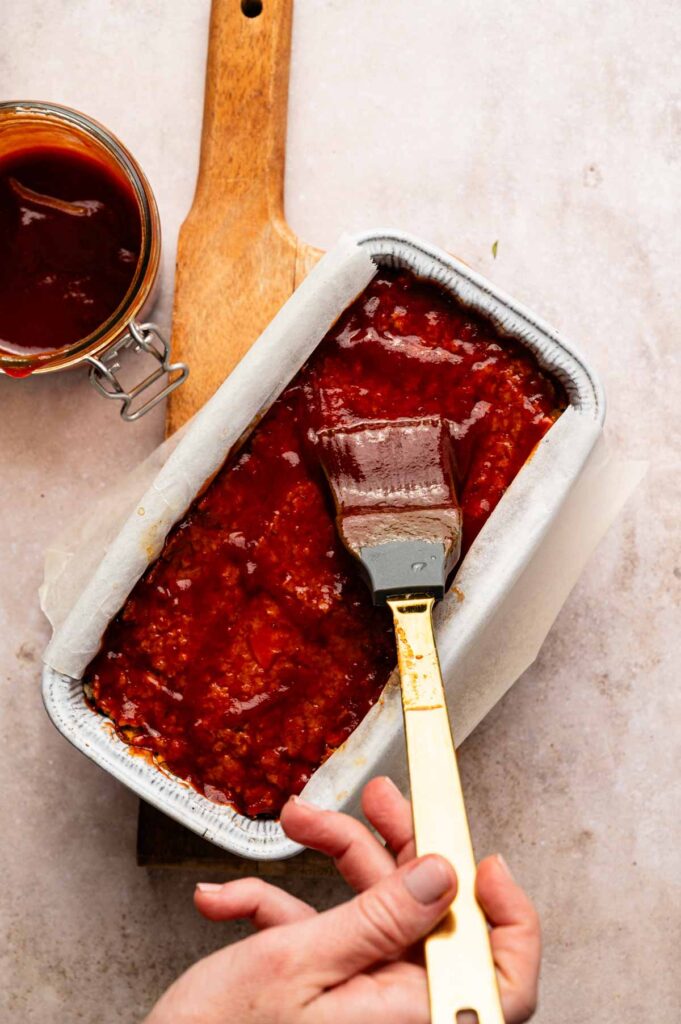 A hand brushes red sauce onto a freshly baked vegetarian meatloaf in a foil pan, with a jar of sauce and wooden cutting board beside it.