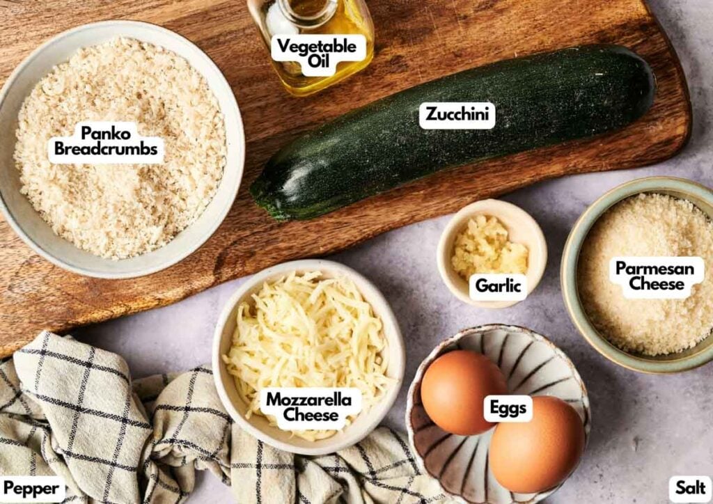 Ingredients for Air Fryer Zucchini Fritters&mdash;including panko breadcrumbs, zucchini, garlic, parmesan cheese, mozzarella, eggs, salt, pepper, and vegetable oil&mdash;are neatly arranged on a countertop.