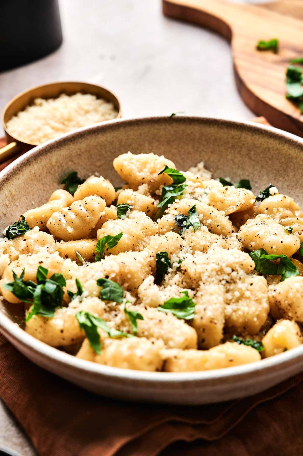 A bowl of Air Fryer Gnocchi topped with grated cheese, black pepper, and fresh herbs, served on a brown cloth with a small dish of grated cheese in the background.