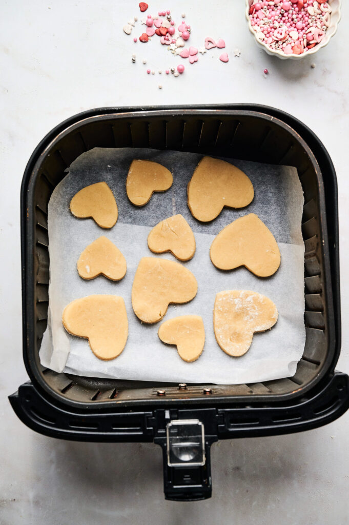 Air Fryer Heart Shaped Cookies: Heart-shaped cookie dough pieces rest on parchment paper inside an air fryer basket, with pink and white sprinkles scattered nearby, ready to be baked into adorable treats.