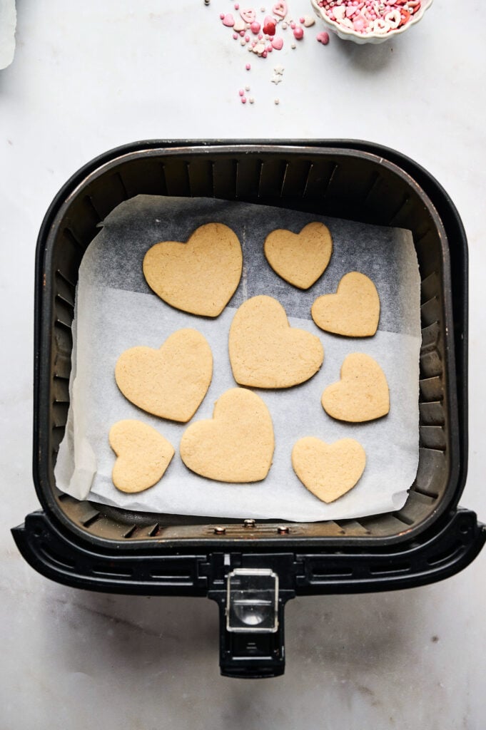 Air Fryer Heart Shaped Cookies are arranged on parchment paper inside an air fryer basket, with a bowl of pink sprinkles visible in the background.