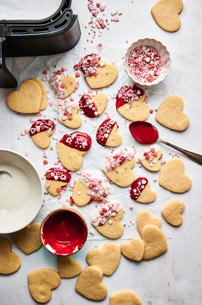 Air Fryer Heart Shaped Cookies, some plain and some decorated with red and white icing and sprinkles, are spread on a parchment-lined surface with small bowls of icing and a spoon nearby.