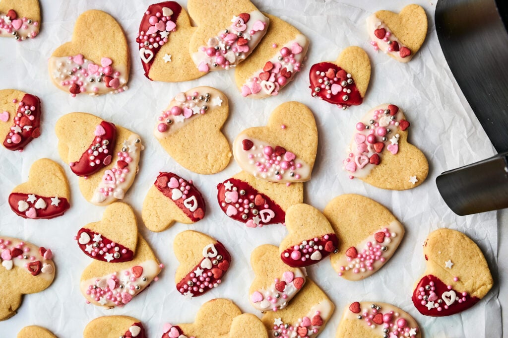 Air Fryer Heart Shaped Cookies decorated with red and white icing and assorted sprinkles are arranged on a sheet of parchment paper.