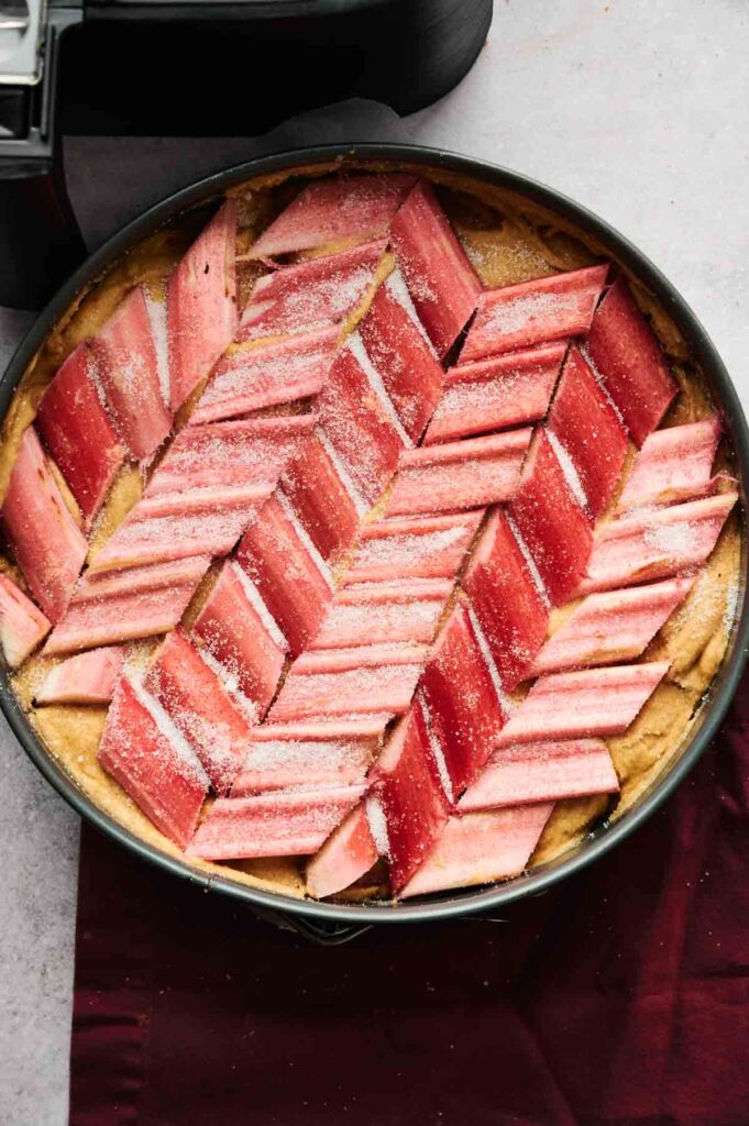 An Air Fryer Rhubarb Cake sits in a round baking dish, its dessert surface topped with rhubarb pieces arranged in a herringbone pattern and lightly dusted with sugar.