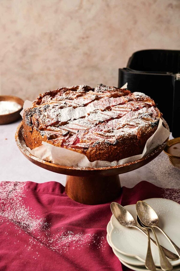 An Air Fryer Rhubarb Cake, freshly baked and dusted with powdered sugar, sits on a wooden stand with a red cloth, plates, and spoons nearby.