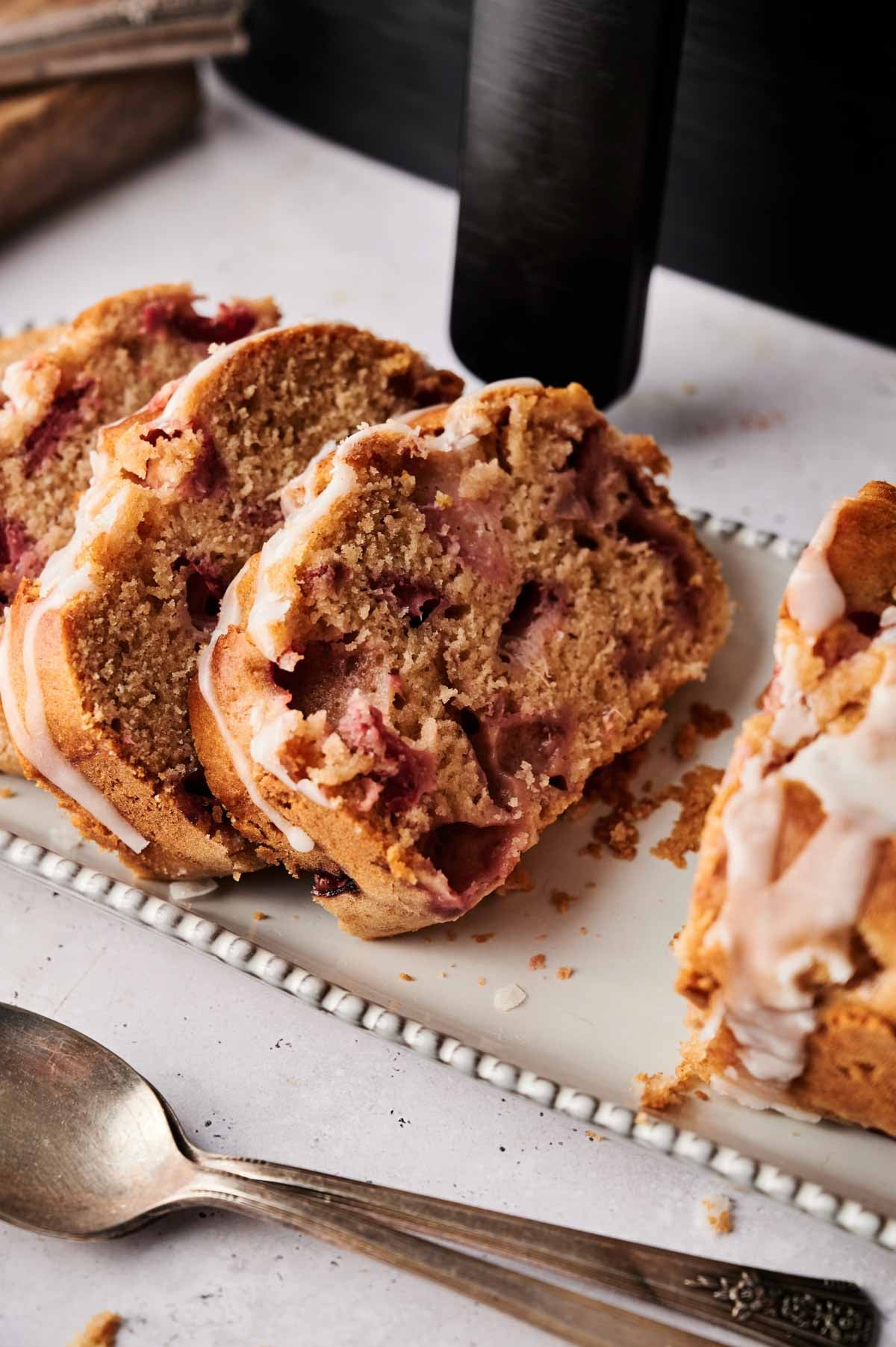 Three slices of Air Fryer Strawberry Bread, glazed to perfection, are arranged on a rectangular white plate next to a vintage spoon.