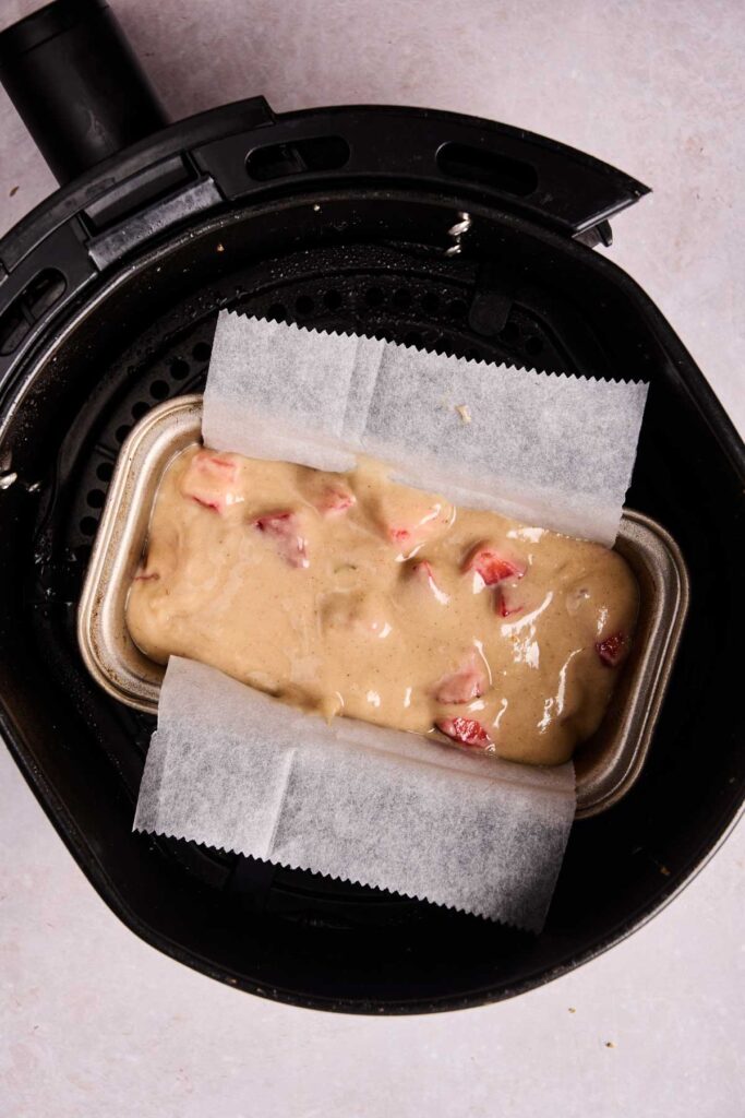 A parchment-lined loaf pan with unbaked Air Fryer Strawberry Bread batter and fresh strawberry pieces sits nestled inside an air fryer basket.