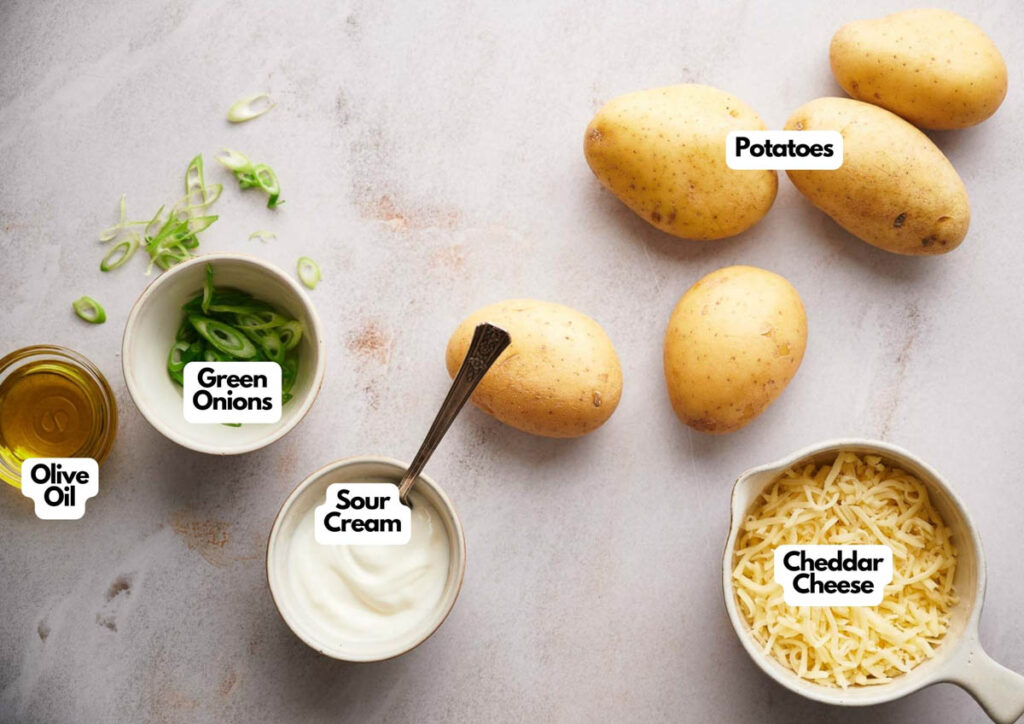 Five potatoes, a bowl of shredded cheddar cheese, a bowl of sour cream with a spoon, sliced green onions, and olive oil on the countertop&mdash;perfect essentials for making crispy Air Fryer Potato Skins at home.