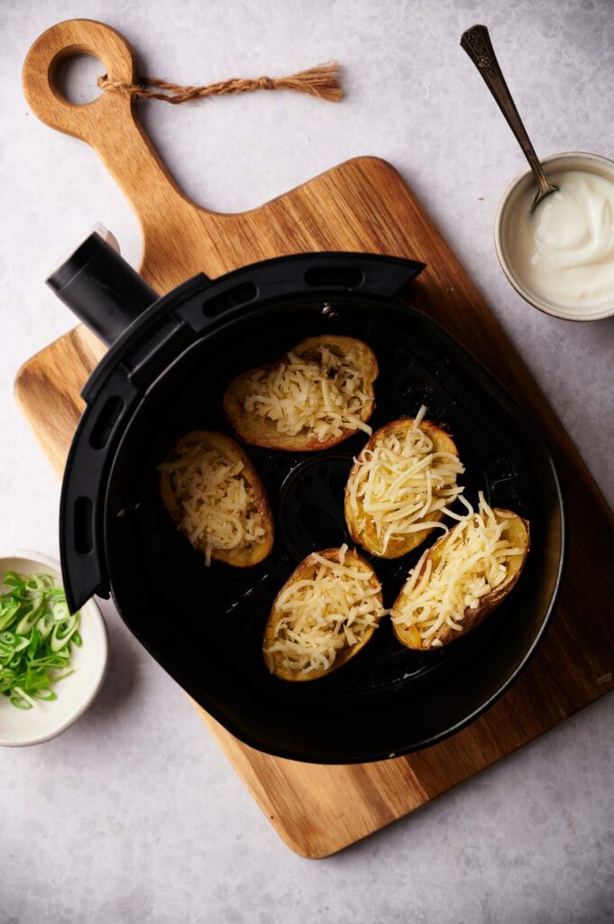 Five Air Fryer Potato Skins filled with shredded cheese rest in a basket on a wooden cutting board, accompanied by bowls of sour cream and chopped green onions for dipping and topping.