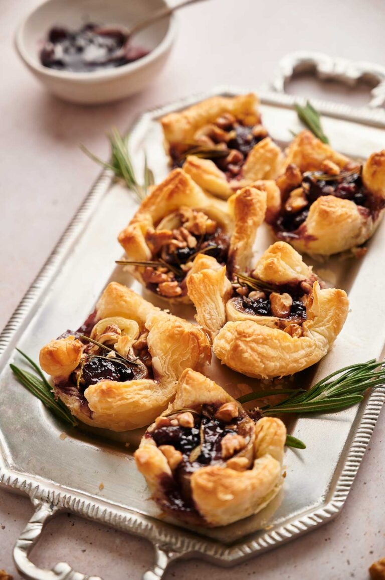 A silver tray holds Cranberry Brie Bites made with puff pastry, dark jam, and nuts, garnished with rosemary sprigs. A bowl with extra jam is in the background.