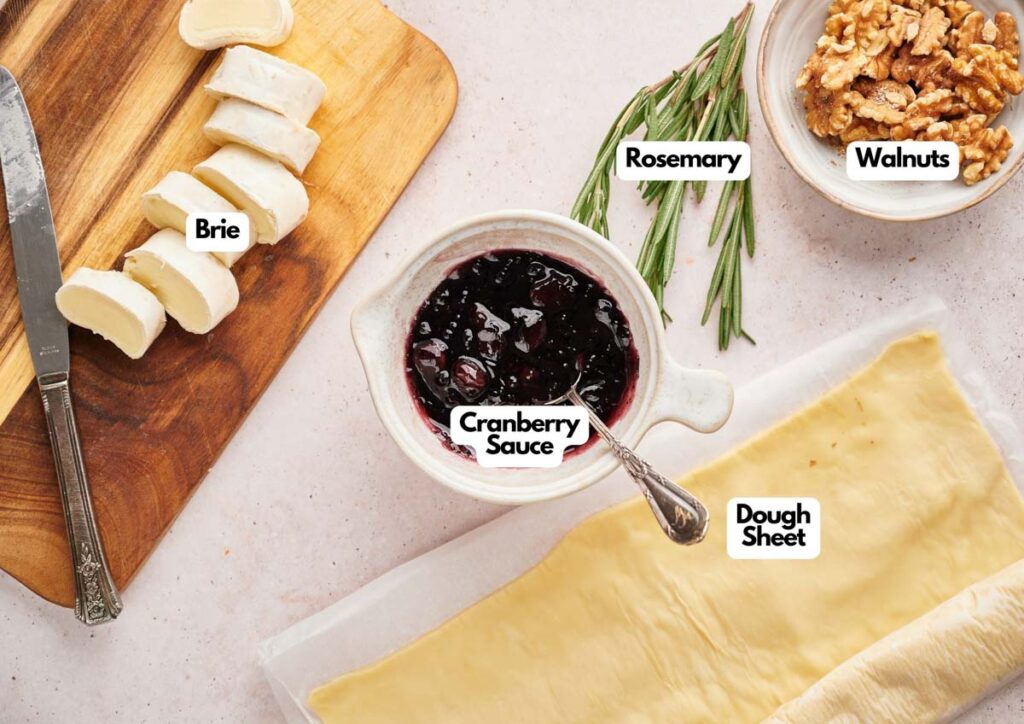 Sliced brie, cranberry sauce, rosemary, walnuts, and a dough sheet are arranged on a light surface, ready for making delicious Cranberry Brie Bites.