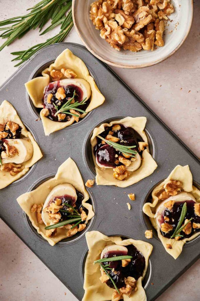 A muffin tin filled with unbaked pastry cups for Cranberry Brie Bites, each holding brie cheese, dark fruit jam, walnuts, and rosemary, with extra walnuts and rosemary on the side.