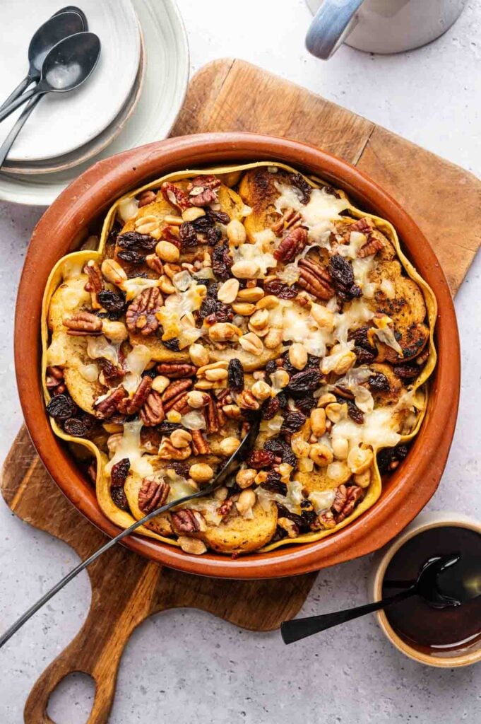 A baked dish in a round ceramic pan holds a delicious Capirotada made with sliced apples, nuts, raisins, and melted cheese. A serving spoon rests in the dish on a wooden board beside plates and utensils.
