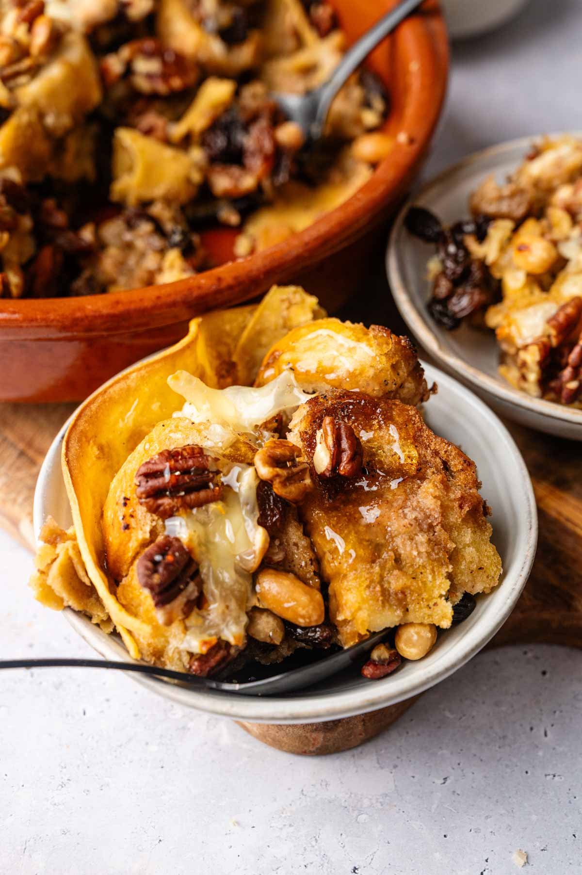 A bowl of capirotada bread pudding topped with mixed nuts and drizzled with honey, with a larger serving dish and another plate in the background.