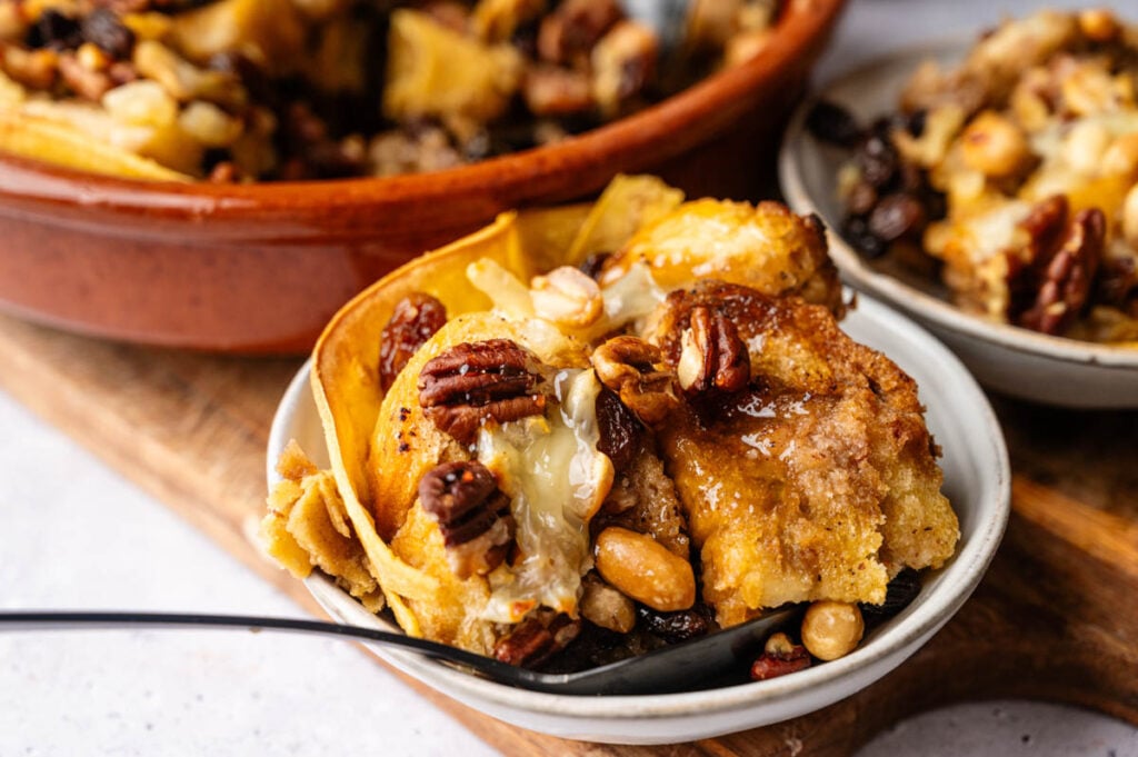 A bowl of capirotada bread pudding topped with mixed nuts and syrup, complete with a spoon inside and a larger serving dish in the background.