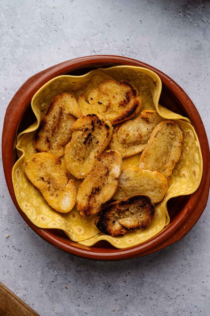 Golden fried bread slices are layered over corn tortillas in a clay dish, the base for capirotada.