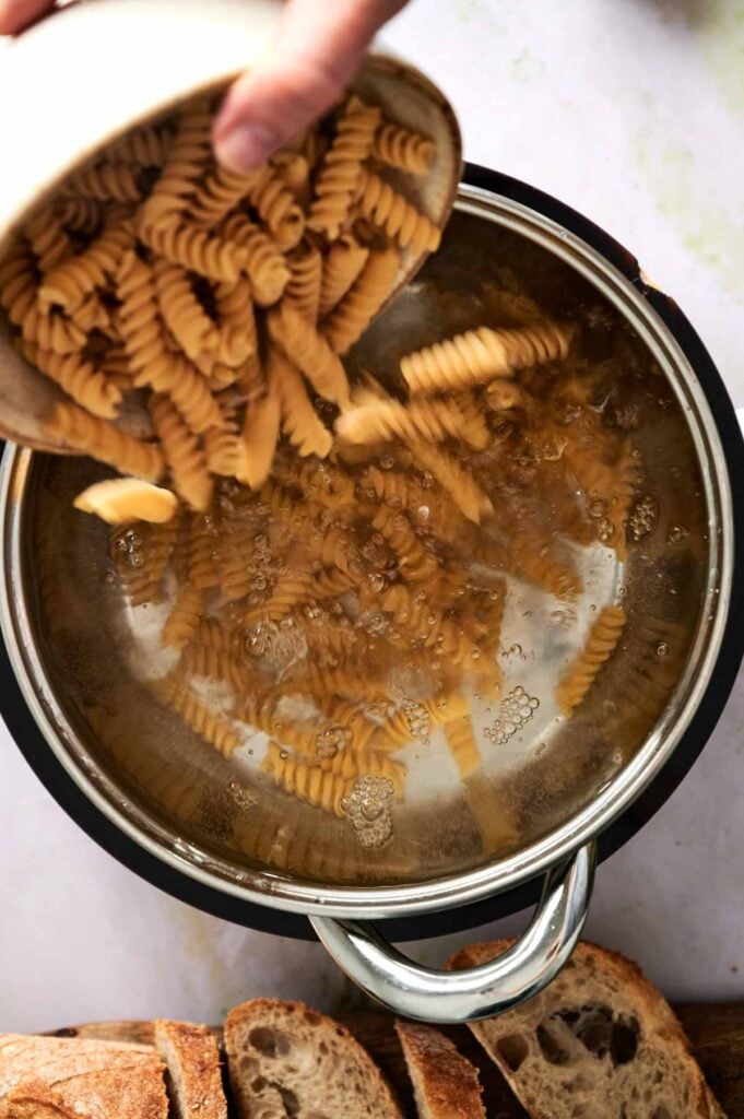 Dry rotini pasta is poured into a pot of boiling water, ready to cook until al dente.