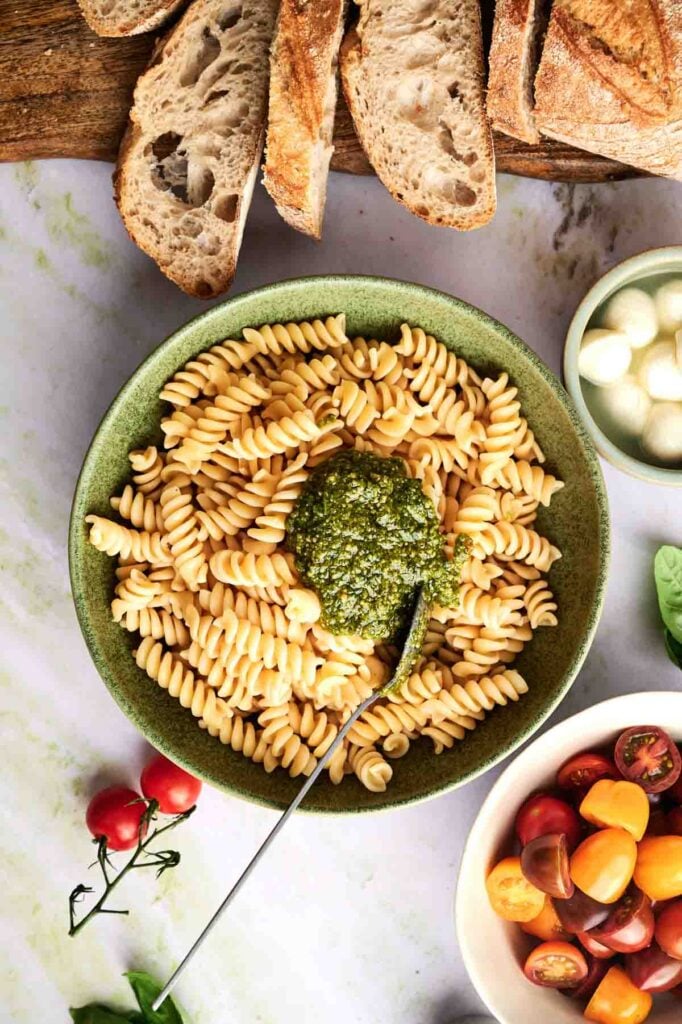 Cooked pasta is topped with bright green pesto in a bowl, with cherry tomatoes and bocconcini nearby.