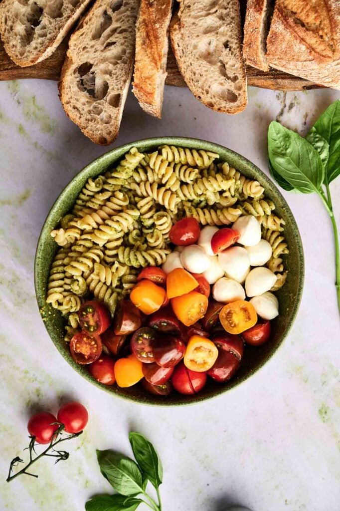 Pesto-coated pasta sits in a bowl next to halved cherry tomatoes and bocconcini, ready to be mixed together.