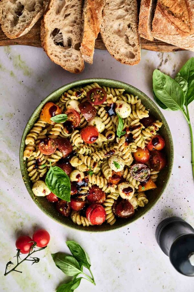 A bowl of pesto pasta salad is topped with fresh basil leaves and balsamic drizzle, ready to serve with crusty bread.