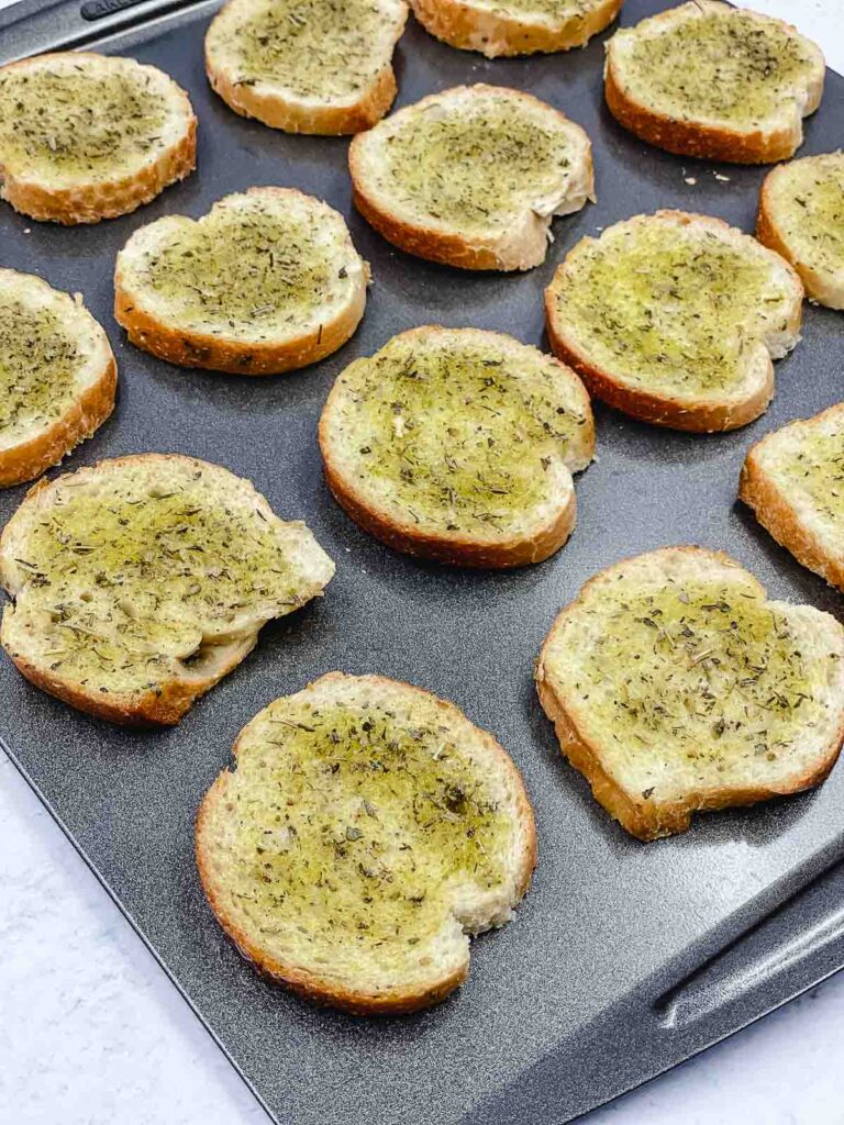 A baking sheet filled with round slices of bread topped with herbs and baked, arranged in rows on the tray, makes for a delightful Caprese appetizer presentation.
