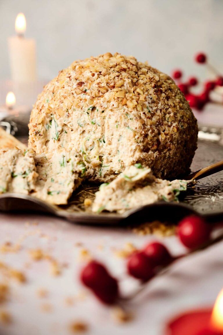 A cheese ball covered in herbs and nuts sits on a silver platter, partially sliced, with scattered walnuts and red berries in the foreground.
