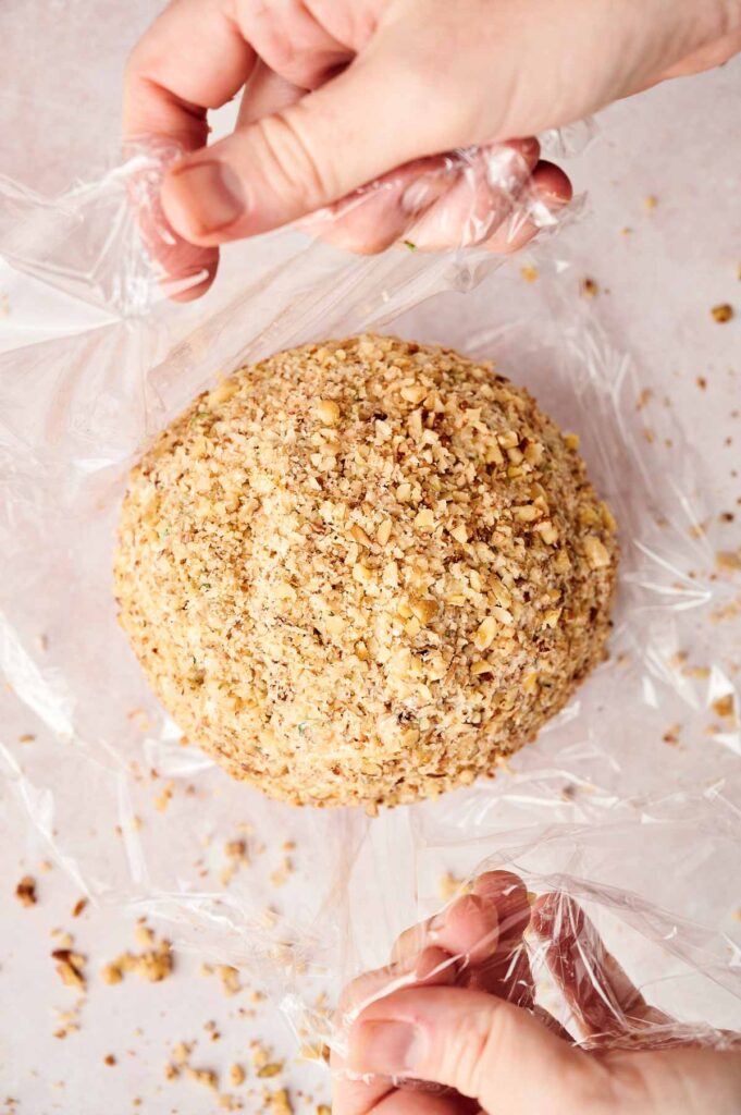 A person's hands are wrapping a cheese ball covered in chopped nuts with plastic wrap.