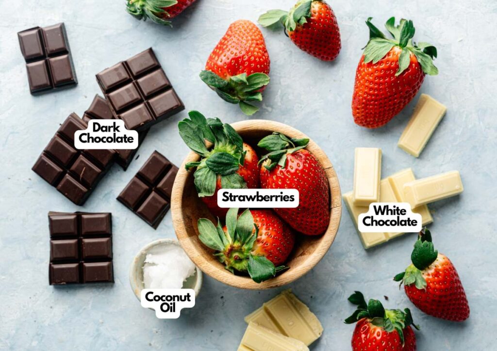 A wooden bowl of strawberries is surrounded by extra strawberries, dark and white chocolate pieces, a small bowl of coconut oil, and labels&mdash;perfect for making delicious chocolate covered strawberries.