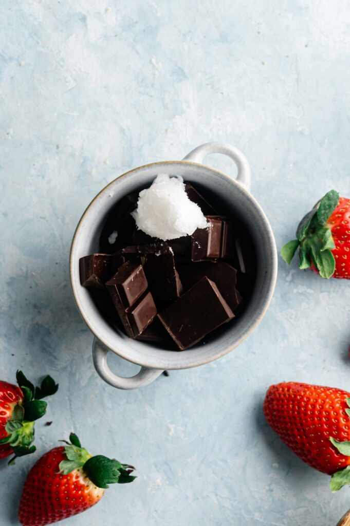 Chopped dark chocolate and a scoop of coconut oil in a small bowl, surrounded by fresh strawberries.