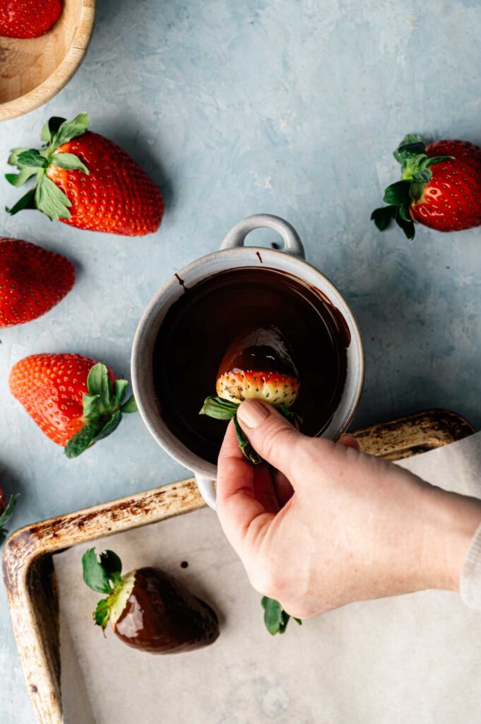 Hand dipping a fresh strawberry into a cup of melted chocolate to make delicious Chocolate Covered Strawberries, with more strawberries and a tray lined with parchment paper nearby.