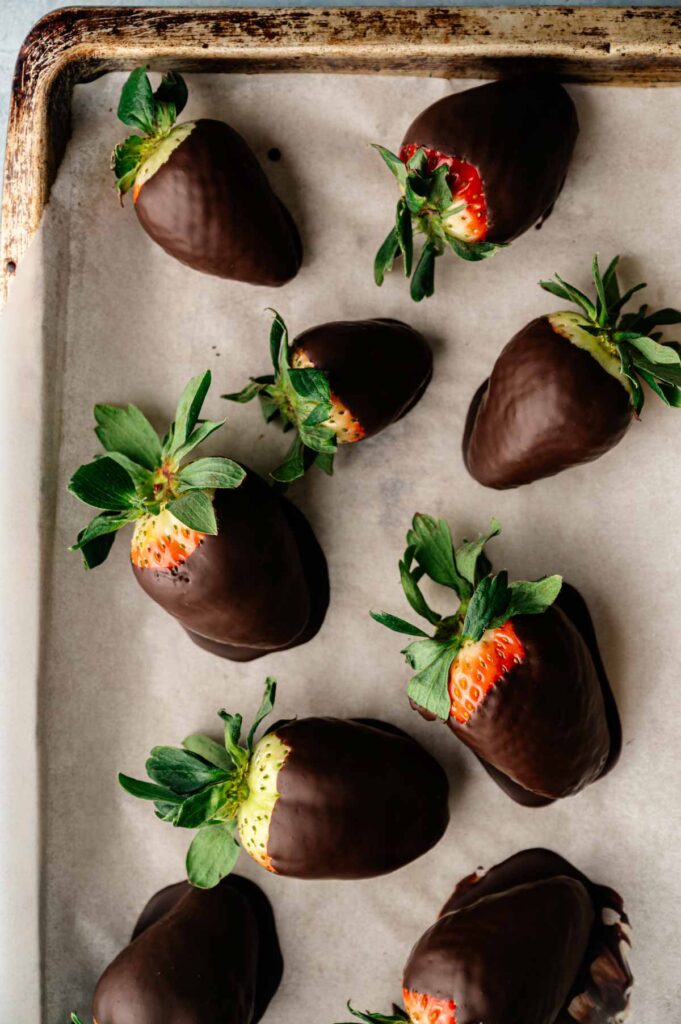 A baking sheet lined with parchment paper holds several chocolate covered strawberries, each berry partially dipped in rich dark chocolate.