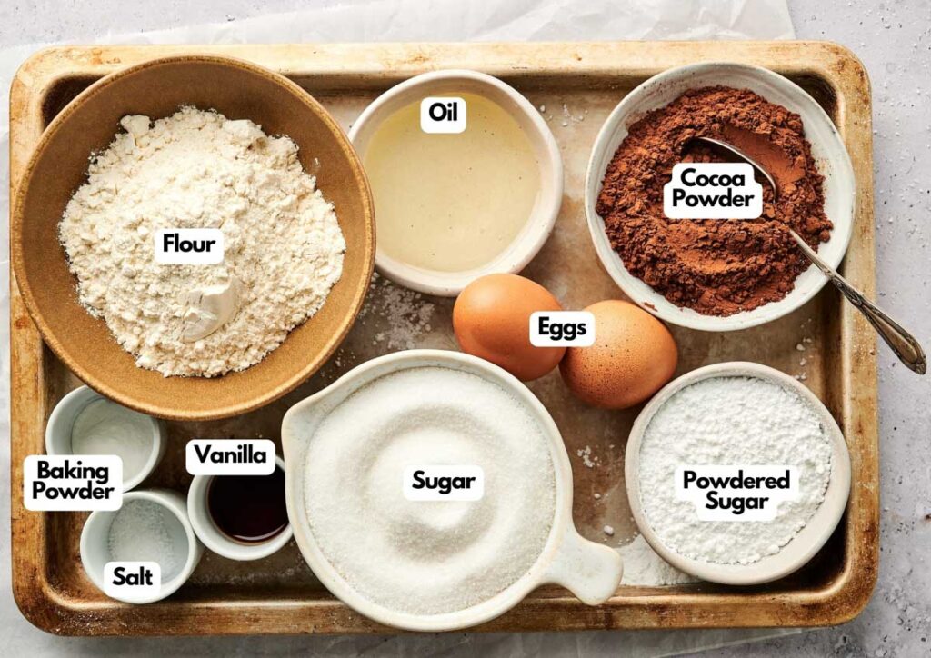A tray with bowls containing flour, cocoa powder, sugar, powdered sugar, baking powder, salt, oil, vanilla extract, and two eggs&mdash;each labeled&mdash;perfectly organized for making Chocolate Crinkle Cookies.