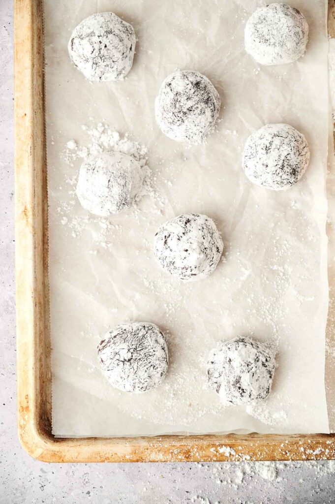 Nine Chocolate Crinkle Cookies dough balls coated in powdered sugar are arranged on a parchment-lined baking sheet.
