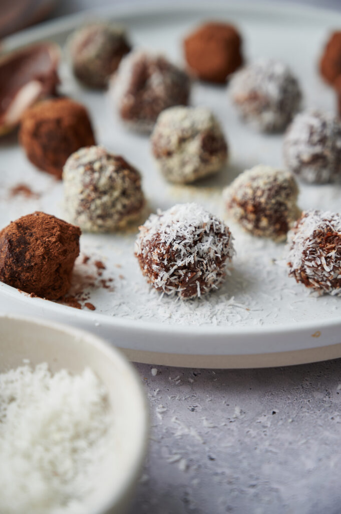 A white plate displays Chocolate Truffles coated in cocoa powder and shredded coconut, with extra coconut flakes in a small bowl in the foreground.
