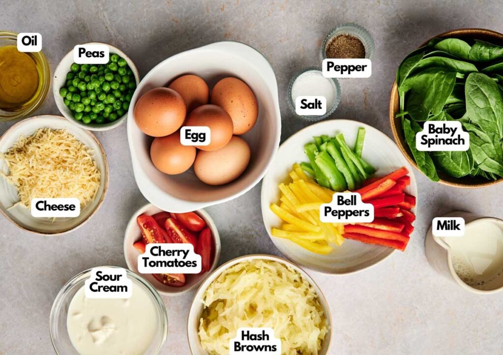 Top-down view of labeled ingredients for an Egg and Potato Breakfast Casserole, featuring eggs, baby spinach, hash browns, bell peppers, peas, cherry tomatoes, cheese, milk, oil, sour cream, salt, and pepper.