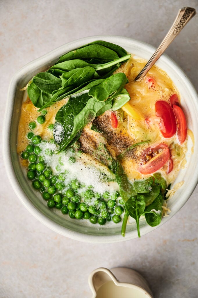 A bowl with whisked eggs, fresh spinach leaves, green peas, sliced tomatoes, grated cheese, and seasoning—with a spoon in the bowl—offers the perfect inspiration for an egg and potato breakfast casserole.