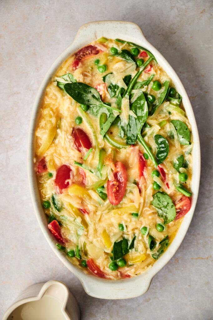 An oval casserole dish filled with a baked pasta dish, reminiscent of an Egg and Potato Breakfast Casserole, featuring shredded cheese, peas, spinach, tomato slices, and yellow bell pepper.