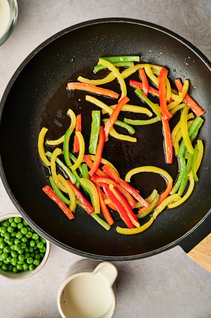 Sliced red, green, and yellow bell peppers sautéing in a black frying pan, with a bowl of green peas and a small jug of cream nearby on a light countertop—perfect colorful ingredients to add into an egg and potato breakfast casserole.
