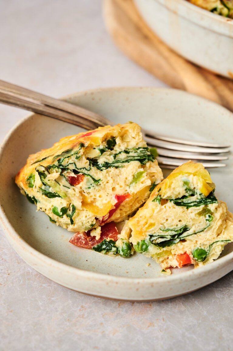Two slices of vegetable frittata, reminiscent of an Egg and Potato Breakfast Casserole, with visible spinach, red bell peppers, and peas served on a beige plate with a fork.