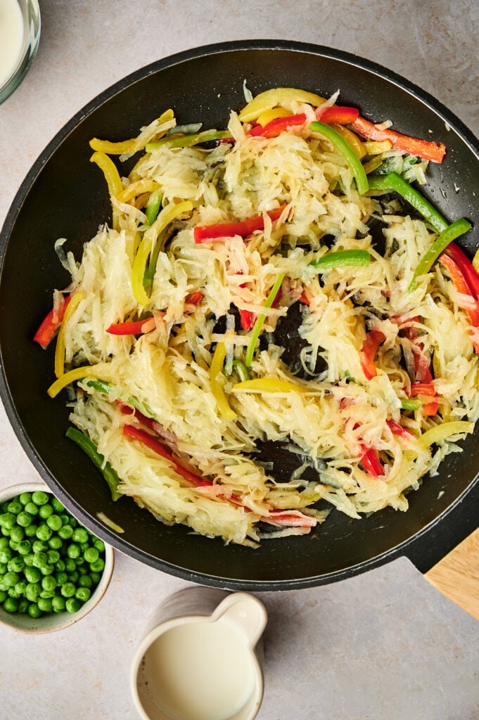 A skillet contains shredded hash browns mixed with red, yellow, and green bell pepper strips, with a small bowl of green peas and a white cup of milk nearby on a light countertop.