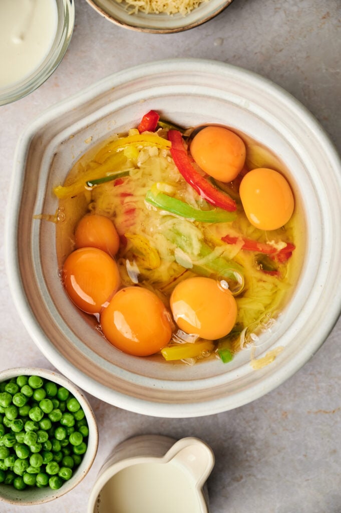 A bowl containing six cracked eggs, shredded hash browns, and sliced bell peppers sits beside bowls of green peas and a small jug of cream, showing the ingredients for an egg and potato breakfast casserole.